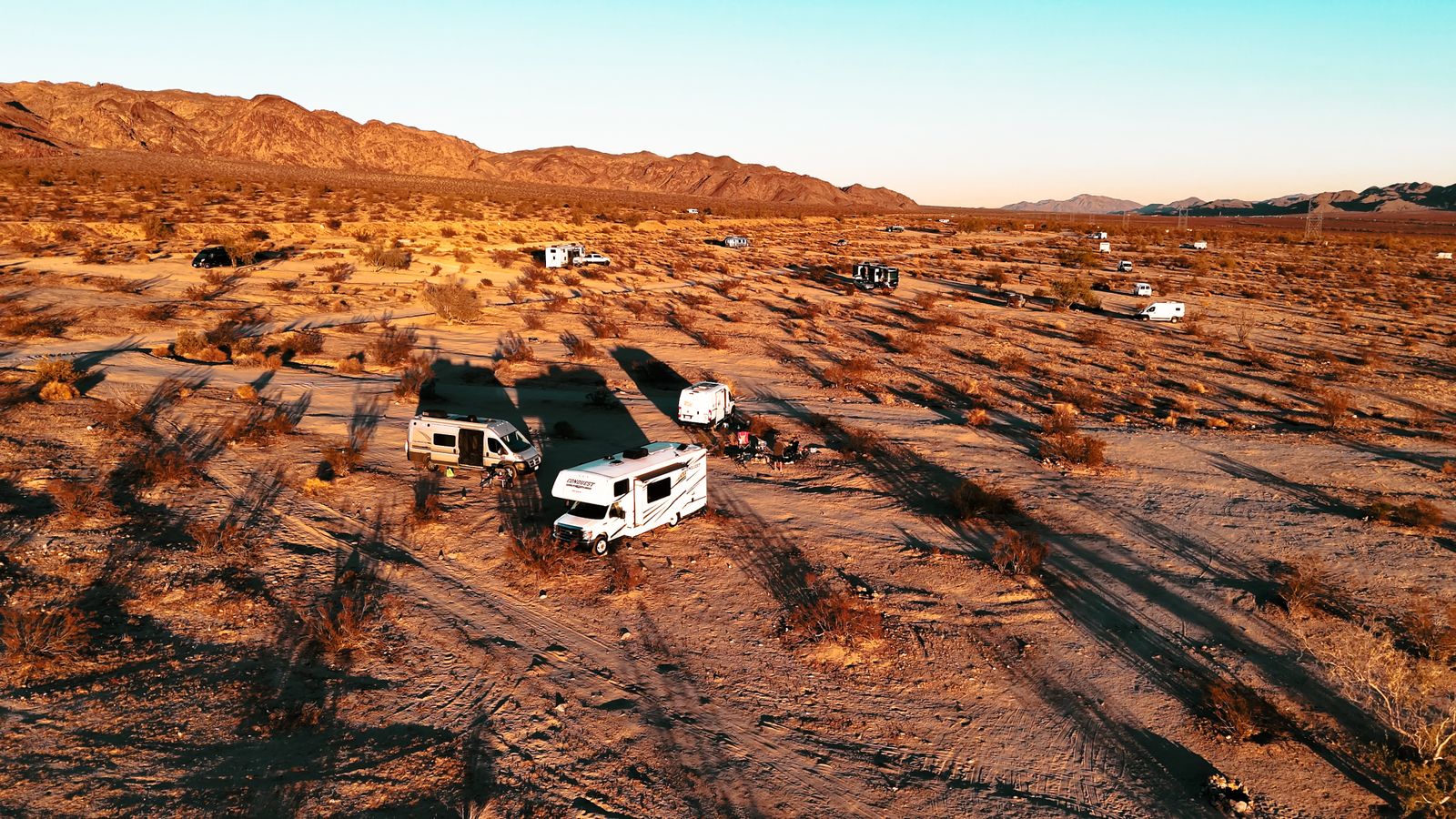 Aerial view of motorhomes and camper vans spread across open desert at sunset with mountains behind