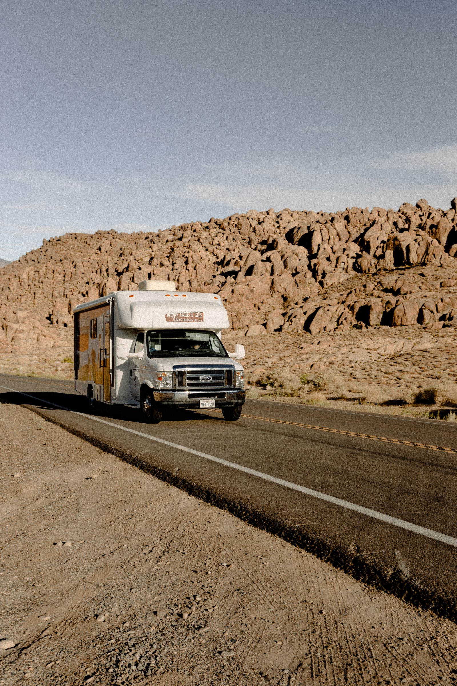 Class C motorhome with a rooftop unit driving a two-lane desert highway past rocky hills
